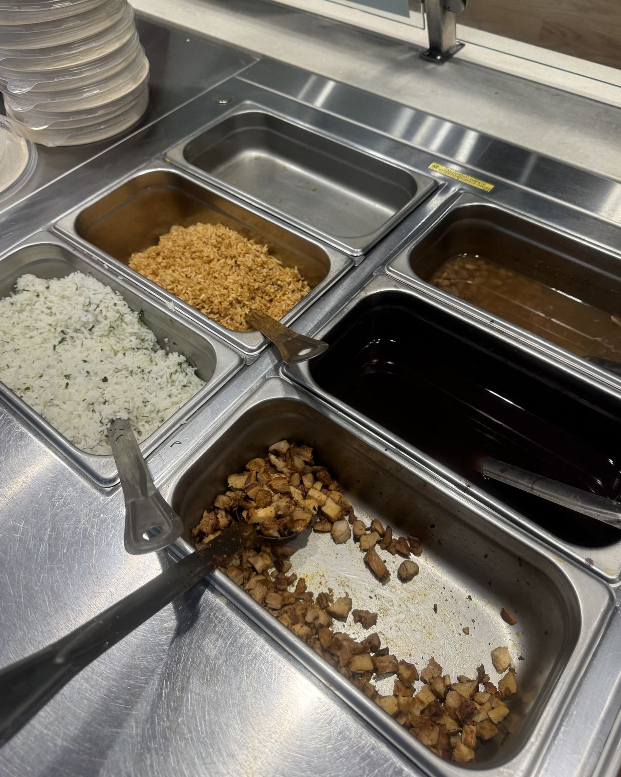 Steel pans on a food line at close: two pans of rice, beans running low, an empty pan, and the last pieces of chicken