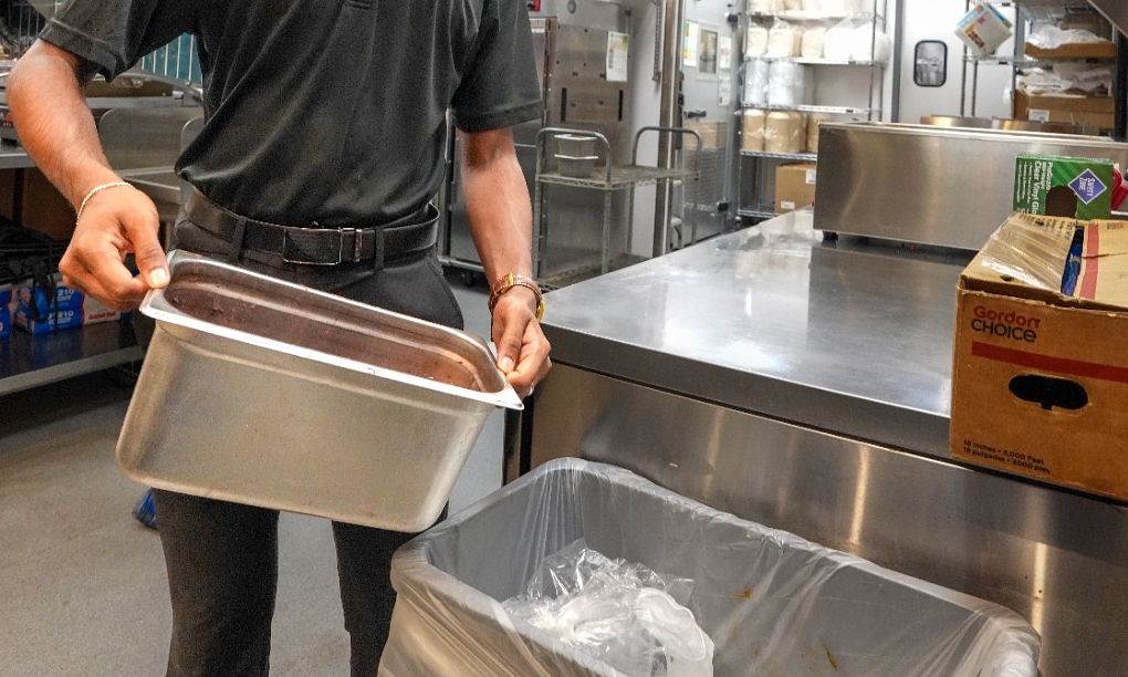 Hands holding a nearly empty steel pan over a lined trash bin in a back kitchen